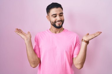 İspanyol genç adam pembe arka planda gülümsüyor. İki elini de açık tutuyor. Karşılaştırma ve reklam karşılaştırması yapıyor ve dengeyi sağlıyor. 