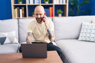 Young bald man talking on smartphone using laptop at home