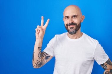 Mavi arka planda dikilmiş, gülümseyen, parmaklarını zafer işareti yaparken gösteren İspanyol dövmeli adam. İki numara.. 