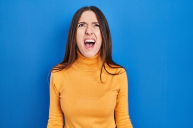 Genç esmer kadın mavi arka planda kızgın ve kızgın bağırıyor sinirli ve öfkeli, öfkeyle bağırıyor. öfke ve agresif konsept. 