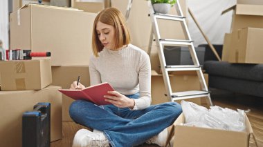 Young blonde woman writing on notebook sitting on floor at new home