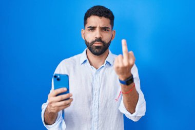 Hispanic man with beard using smartphone typing message showing middle finger, impolite and rude fuck off expression 