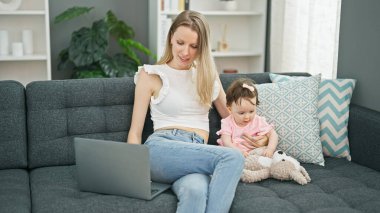 Anne ve kızı kanepede oturup oyuncak ayıyla oynuyorlar. Evde dizüstü bilgisayar kullanıyorlar.