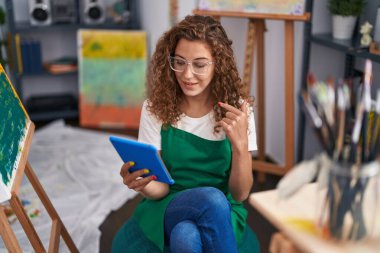 Beyaz kadın ressam, resim stüdyosunda oturmuş, tabletle video görüşmesi yapıyor, mutlu bir şekilde el ve parmak işareti yapıyor. 
