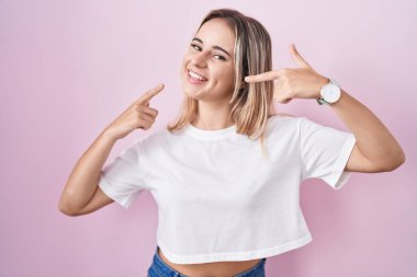 Young blonde woman standing over pink background smiling cheerful showing and pointing with fingers teeth and mouth. dental health concept. 