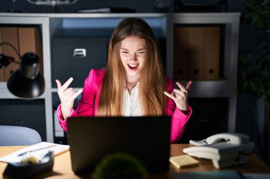 Young caucasian woman working at the office at night shouting with crazy expression doing rock symbol with hands up. music star. heavy concept. 