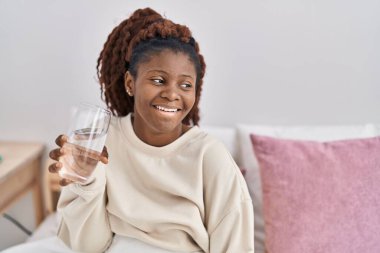 African american woman drinking glass of water sitting on bed at bedroom