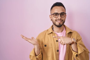 Young hispanic man standing over pink background amazed and smiling to the camera while presenting with hand and pointing with finger. 