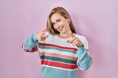 Young blonde woman standing over pink background pointing fingers to camera with happy and funny face. good energy and vibes. 