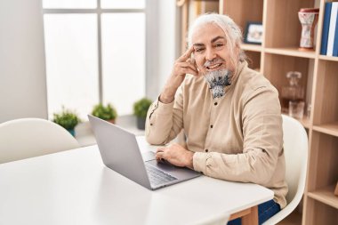 Evdeki masada oturan orta yaşlı, kır saçlı, dizüstü bilgisayar kullanan bir adam.