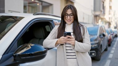 Young beautiful hispanic woman using smartphone leaning on car at street