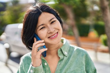 Genç Çinli kadın gülümsüyor. Parktaki akıllı telefondan konuşuyor.