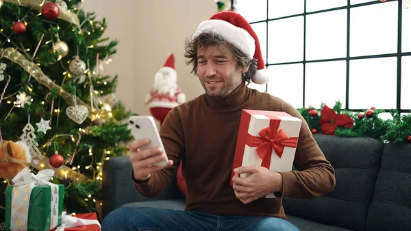 自宅でクリスマスプレゼントを持ってソファーに座ってスマートフォンを使用する若いヒスパニック男性 — ストック写真