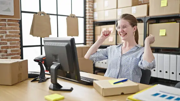 Young blonde woman ecommerce business worker using computer writing on package with winner expression at office
