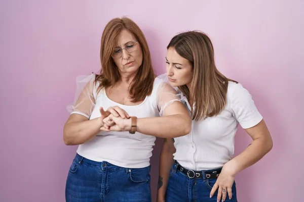 Hispanic mother and daughter wearing casual white t shirt over pink background checking the time on wrist watch, relaxed and confident