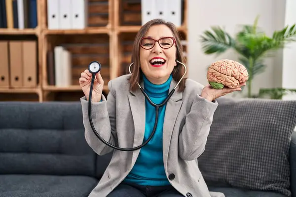Middle age hispanic woman working at therapy office holding brain smiling and laughing hard out loud because funny crazy joke.