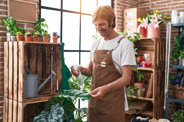 Young blond man florist watering plant working at flower shop