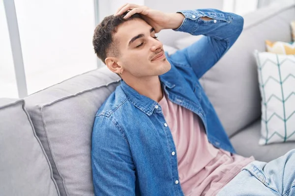 Young hispanic man relaxed with hand on head sitting on sofa at home
