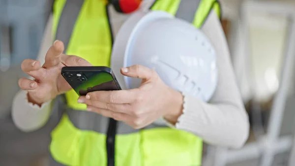 Young blonde woman architect using smartphone holding hardhat at construction site
