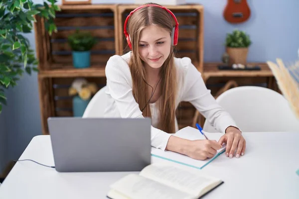 Young caucasian woman writing on notebook listening to music studying at home