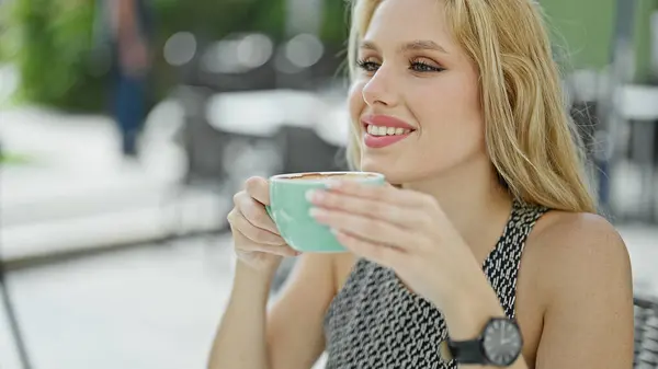 Young blonde woman smelling cup of coffee sitting on table at coffee shop terrace