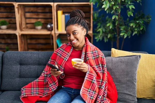 Afro-Amerikalı kadın kahve içiyor ve evde akıllı telefon kullanıyor.