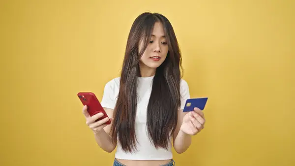 Young chinese woman shopping with smartphone and credit card with relaxed expression over isolated yellow background