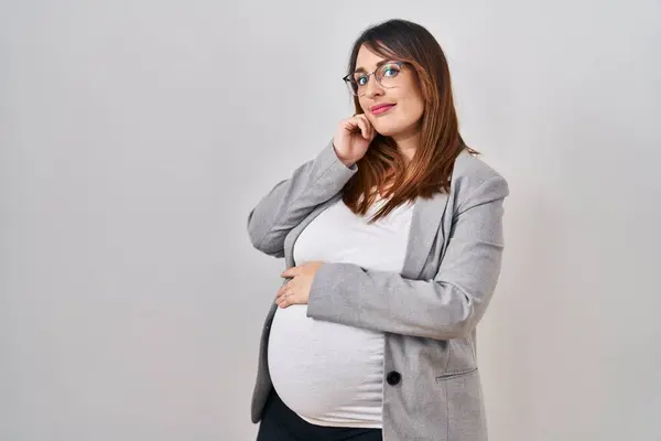 怀孕的女商人站在白种人的背景上严肃地思考着手托着下巴的问题 思考着令人困惑的想法 — 图库照片