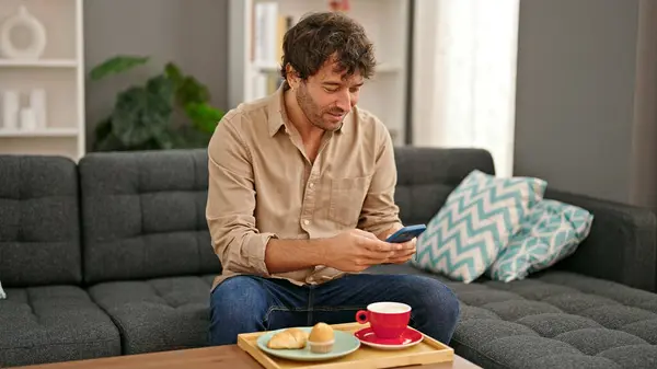 Young hispanic man having breakfast using smartphone smiling at home