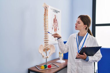 Genç sarışın kadın pisiyoterapist rehabilitasyon kliniğinde elinde pano tutan omurların anatomik modelini gösteriyor.