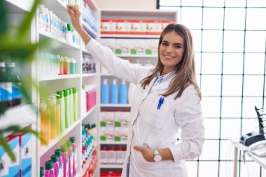 Genç ve güzel İspanyol kadın eczacı gülümseyerek eczanede kendine güvenen ürünleri raflarda tutuyor.