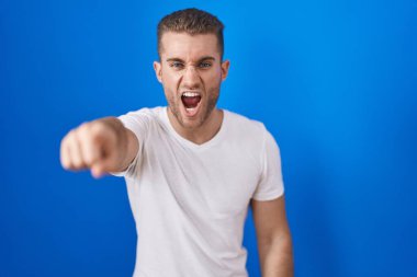 Mavi arka planda dikilen genç beyaz adam kameraya kızgın ve sinirli olduğunu gösteriyor. 