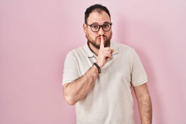 Artı, pembe arka planda duran sakallı, dudakları parmaklı, sessiz olmak isteyen İspanyol bir adam. sessizlik ve gizli konsept. 