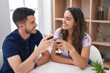 Young hispanic couple drinking wine sitting on table at home