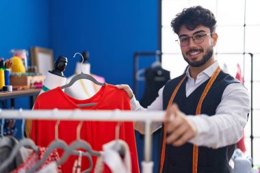 Young hispanic man tailor smiling confident holding clothes on rack at sewing studio