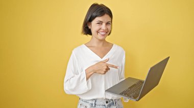 Genç, güzel, İspanyol bir kadın, sırıtarak dizüstü bilgisayara bakarak izole edilmiş sarı arka planı işaret ediyor.