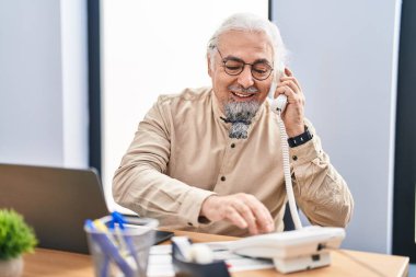 Middle age grey-haired man business worker talking on telephone using laptop at office