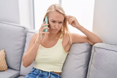 Evde ciddi bir ifadeyle akıllı telefondan konuşan genç sarışın kadın.