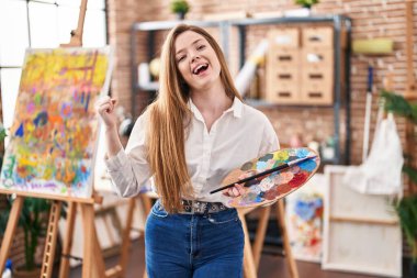 Sanat stüdyosundaki genç beyaz kadın palet tutuyor gururla bağırıyor zaferi kutluyor ve başarıyı kollarını kaldırarak kutluyor. 