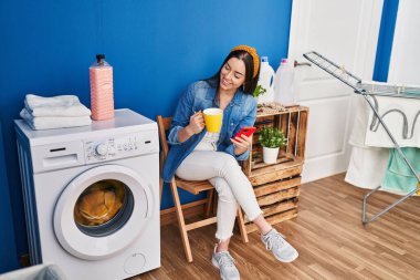 Genç ve güzel İspanyol kadın akıllı telefon kullanıyor kahve içiyor çamaşır odasında çamaşır makinesini bekliyor.