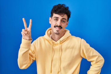Hispanic man standing over blue background smiling looking to the camera showing fingers doing victory sign. number two. 
