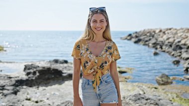 Young blonde woman smiling confident standing at seaside