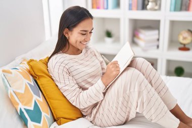 Young beautiful hispanic woman writing on notebook sitting on bed at bedroom