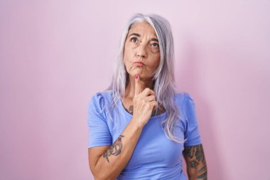 Pembe arka planda duran orta yaşlı bir kadın şüpheye odaklanmış, çenesinde parmak olan ve kafasını kaldırıp merak eden. 