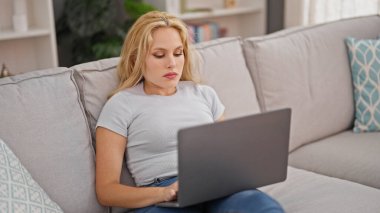 Evdeki kanepede dizüstü bilgisayar kullanan genç sarışın kadın.