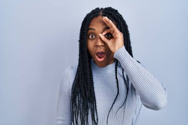 Mavi arka planda dikilen Afro-Amerikalı kadın şaşırmış yüz ifadesiyle şaşırmış, parmaklarının arasından bakan bir jest yapıyor. İnançsız ifade. 