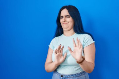 Mavi arka planda duran mavi saçlı genç modern kız tiksindirici ve tiksindirici bir ifade takınıyor çünkü tiksindirici bir tepki veriyor.. 