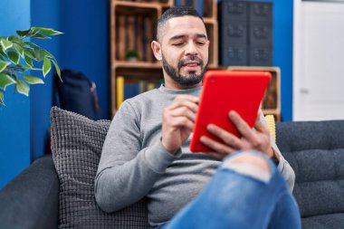 Evdeki koltukta oturan dokunmatik ped kullanan genç latin adam.