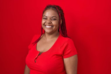 Saçları örgülü, kırmızı arka planda duran, kameraya seksi bir ifadeyle bakan, neşeli ve mutlu yüzlü Afro-Amerikan kadın.. 