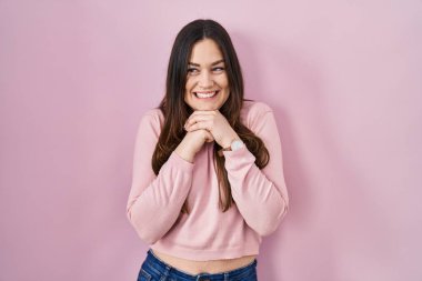 Young brunette woman standing over pink background laughing nervous and excited with hands on chin looking to the side 